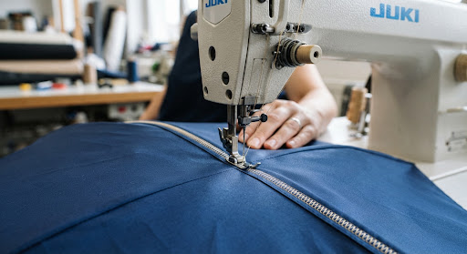 Close-up of a precision industrial sewing machine stitching a high-quality umbrella canopy. The focus is on the tight, uniform stitching and the vibrant color of the fabric. The lighting is sharp, accentuating the mechanical detail and the craftsmanship involved in high-end umbrella production.