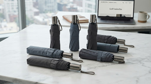 An elegant, slim-profile fashion umbrella with a handcrafted maple wood handle and a deep navy canopy. The image is shot in a luxurious, minimalist urban setting with a soft focus on the rain-slicked pavement. Lighting is sophisticated and moody, emphasizing the texture of the fabric and the polished finish of the wood. The aesthetic is clean, timeless, and tailored for luxury retail.